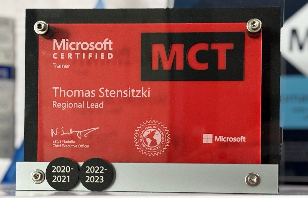 Microsoft Certified Trainer (MCT) Auszeichnung in Rot und Schwarz, mit dem Namen „Thomas Stensitzki“ und der Rolle „Regional Lead“. Sichtbar sind das Microsoft-Logo, die Unterschrift von Satya Nadella, sowie zwei runde Plaketten mit den Jahresangaben „2020–2021“ und „2022–2023“. Die Auszeichnung ist in einem transparenten Rahmen mit Metallschrauben befestigt.